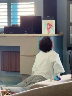 Человек лежит на животе у комода в комнате рядом с рабочей зоной; видны картина, колонки и лампа — бытовая повседневная сцена.