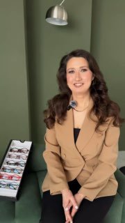 Дарья в бежевом пиджаке сидит на зелёном фоне и улыбается; слева выставочный стенд с оправами O.D.L., включая Azart.