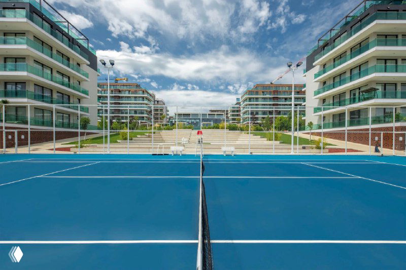 Теннисный корт внутри закрытого жилого комплекса: вид на корты в центре двора и многоэтажные здания по бокам при ясном небе.