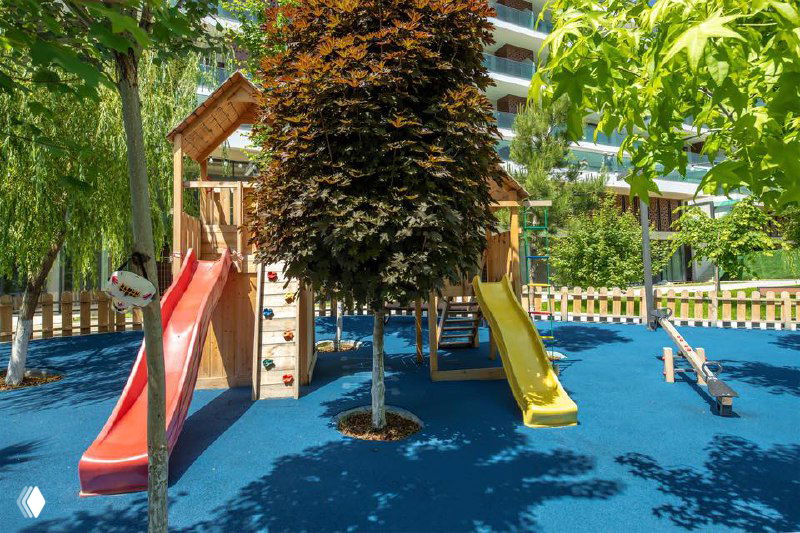 Детская игровая площадка в закрытом комплексе: горки, мягкое покрытие и зелёные деревья вокруг, безопасная зона для детей.