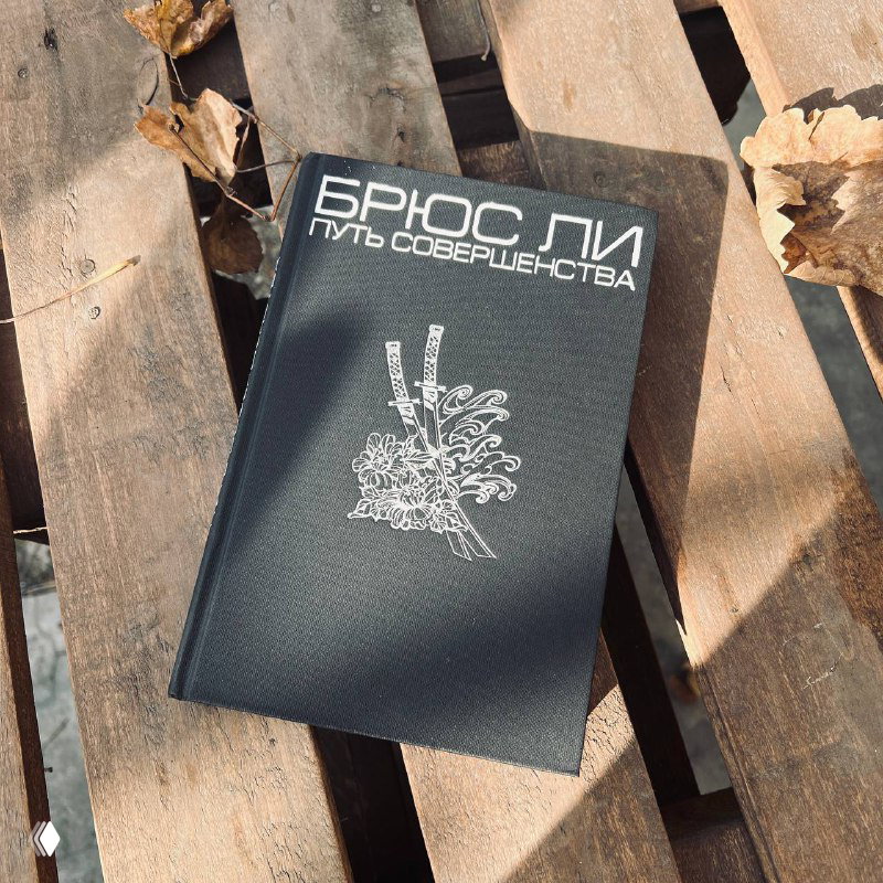 Чёрная обложка книги «Брюс Ли. Путь совершенства» на деревянной поверхности рядом с осенними листьями; крупный план обложки.