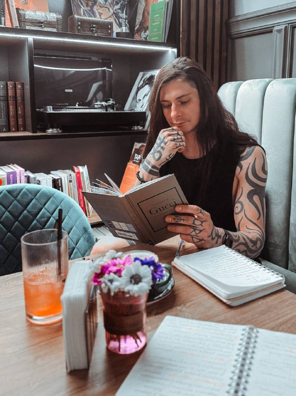 Женщина в кафе переворачивает страницу и читает книгу, перед ней открыт блокнот с заметками и бокал напитка на столе.