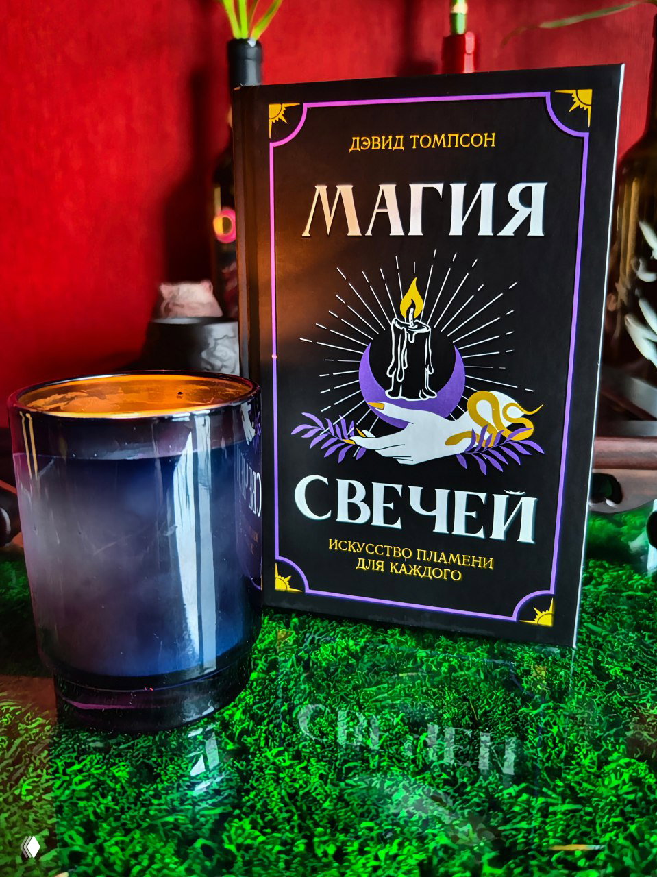 Книга «Магия свечей» рядом с затемнённой стеклянной свечой на столе; обложка с иллюстрацией горящей свечи, луны и рук.