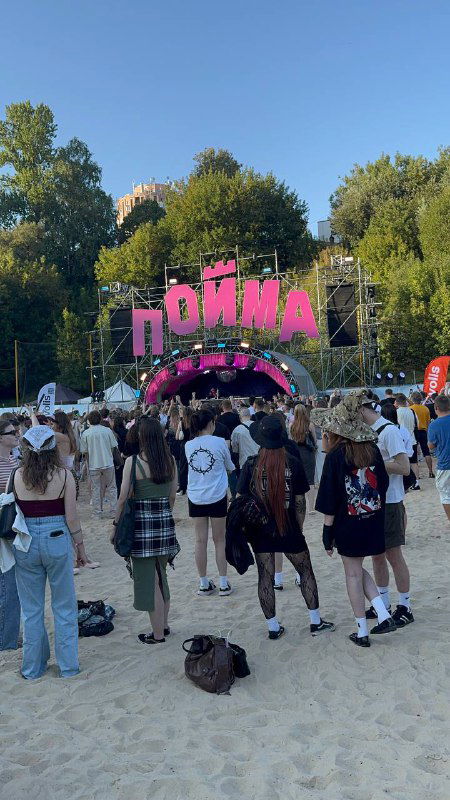 Входная зона фестиваля «Пойма»: большая розовая арка с надписью «Пойма», толпа молодых людей на песчаной площадке и летняя атмосфера перед выступлениями.