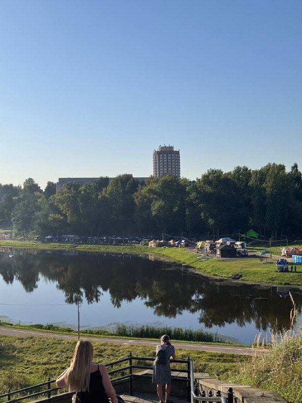 Панорама береговой зоны фестиваля у озера: отдыхующие на траве и берегу, палатки и сцены на фоне зелёных деревьев и высотного дома вдали, тихий летний пейзаж.