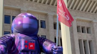 Фигурка талисмана PHDays в фиолетовом скафандре на фоне фасада и флагов фестиваля у входа в Лужники, атмосфера подготовки к событию.