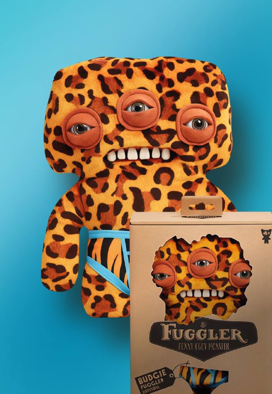 Квадратная плюшевая игрушка Fuggler в леопардовом принте с тремя глазами и открытой пастью рядом с коробкой на однотонном фоне