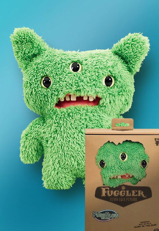 Зелёная плюшевая игрушка Fuggler с тремя глазами и зубами, игрушка стоит рядом с коробкой, фон нейтральный бирюзовый