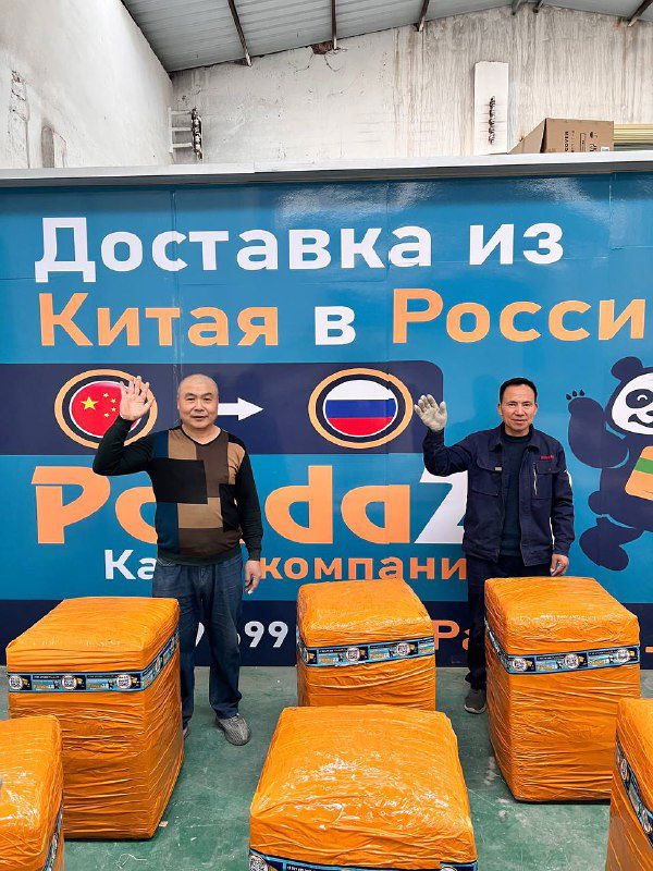 Два сотрудника позируют возле нескольких крупно упакованных посылок в оранжевой защитной плёнке на фоне фирменного баннера.