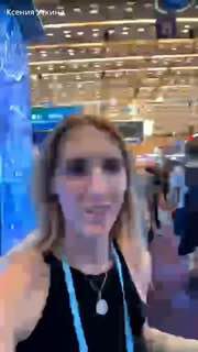 Крупный план ведущей в режиме live на Кантонской выставке: выставочный зал, стенды и посетители на фоне экспонатов и инсталляций.