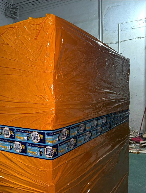 Высокая стопка паллет, обернутая оранжевой плёнкой и зафиксированная лентой Panda28; кадр показывает складскую готовность к отгрузке.