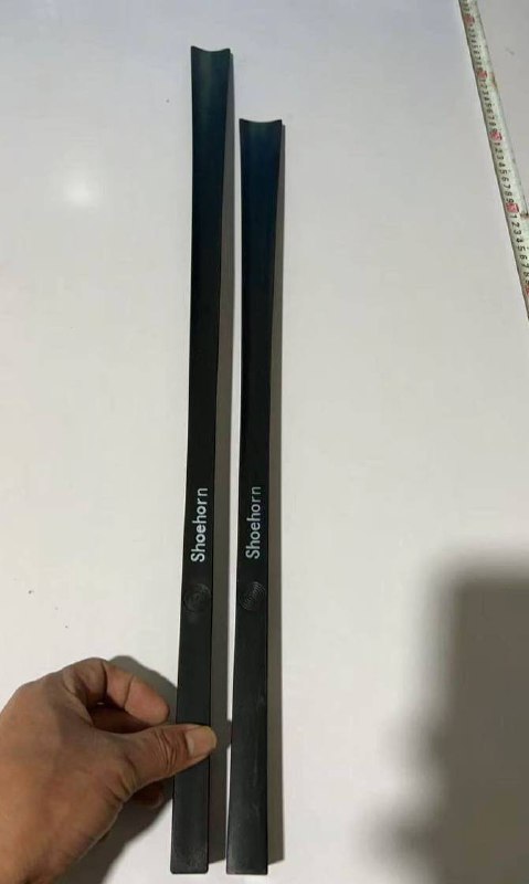 Две чёрные ложки для обуви на белой поверхности рядом с рукой оператора; видна форма изделий и маркировка, фото для контроля качества.