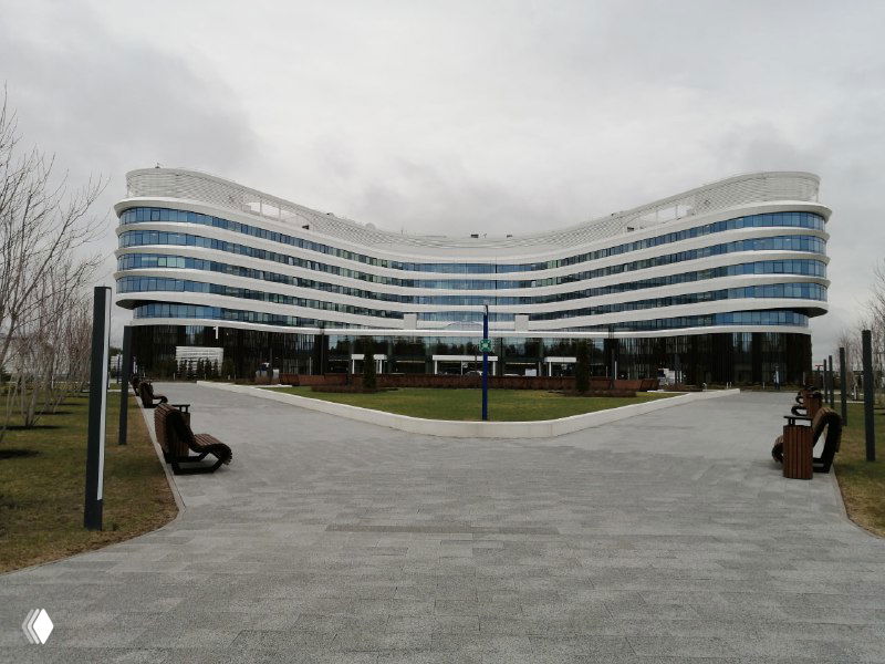 Фронтальный панорамный вид на волнообразный стеклянный фасад клиники Белоостров и входную зону с дорожкой и лавочками.