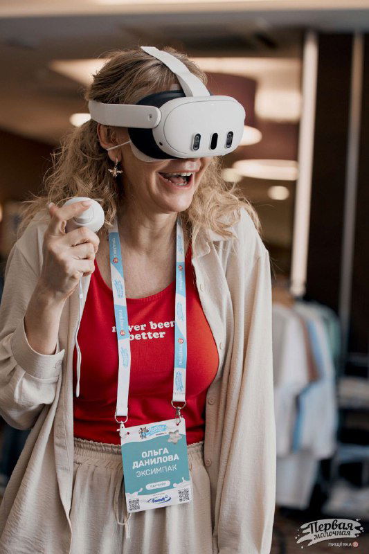 Посетительница в VR-гарнитуре и бейджем на конференции, на заднем плане стенды и другие участники, демонстрация технологии и взаимодействие.