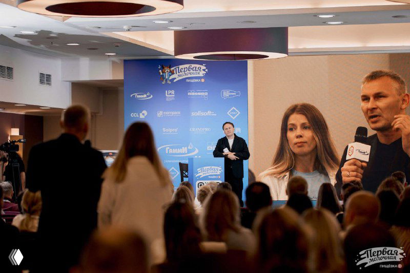 Спикер выступает перед аудиторией, крупный экран показывает лица гостей и брендовые баннеры конференции «Первая Молочная», зал полон слушателей.