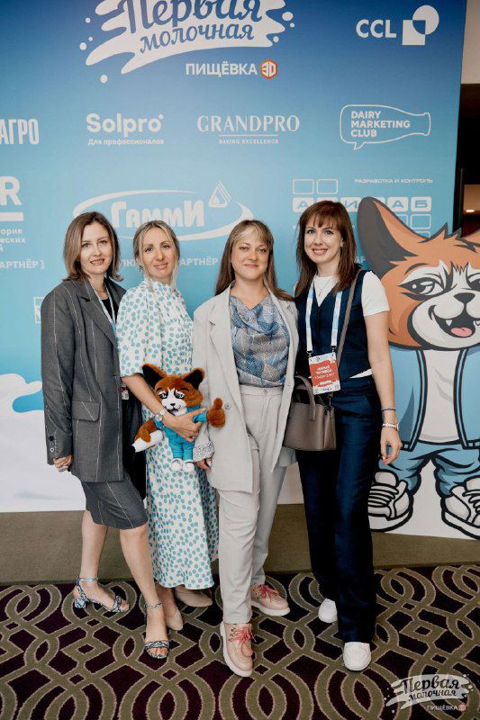 Группа участниц у брендволла с маскотом «Первая Молочная», участники с бейджами, фотография в фотозоне на фоне логотипов партнёров.