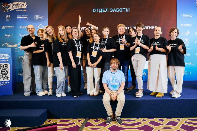 Команда организаторов и волонтёров на сцене мероприятия в унифицированных футболках, групповое фото на фоне баннера спонсоров и логотипа конференции.