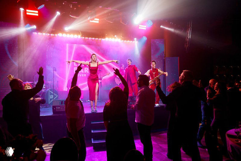 Артистка выступает на сцене перед публикой: яркая подсветка, танцевальный номер и люди, поднимающие руки в такт музыке, вечер в клубной атмосфере.