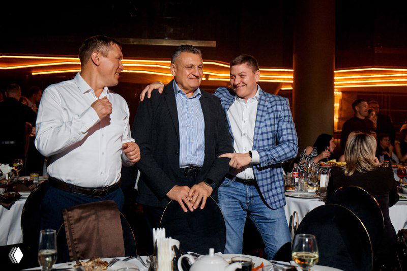 Три гостя вечера стоят у стола и общаются в праздничной атмосфере: деловые беседы за ужином на фоне освещённого банкетного зала Meat Night.