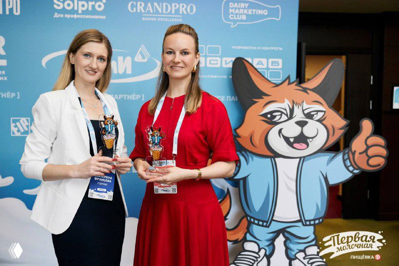 Две женщины с призами на фоне бренд-стены и ростовой фигурой маскота, момент награждения или презентации сотрудничества «Гамми» с отраслевыми партнёрами.