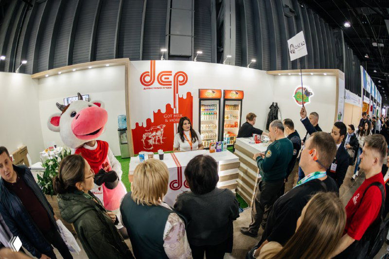 Панорамный кадр одного из стендов выставки с маскотом коровы и многолюдной зоной демонстрации продуктов и упаковки для посетителей.