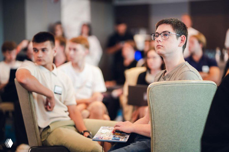 Close-up of attentive young participants in the audience, some holding notebooks and conference materials while focusing on the talk and slides.