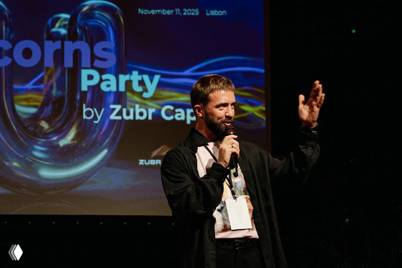Speaker on stage holding a microphone during the Future Unicorns Party 2025 in Lisbon, branded backdrop showing event title and sponsor logos.