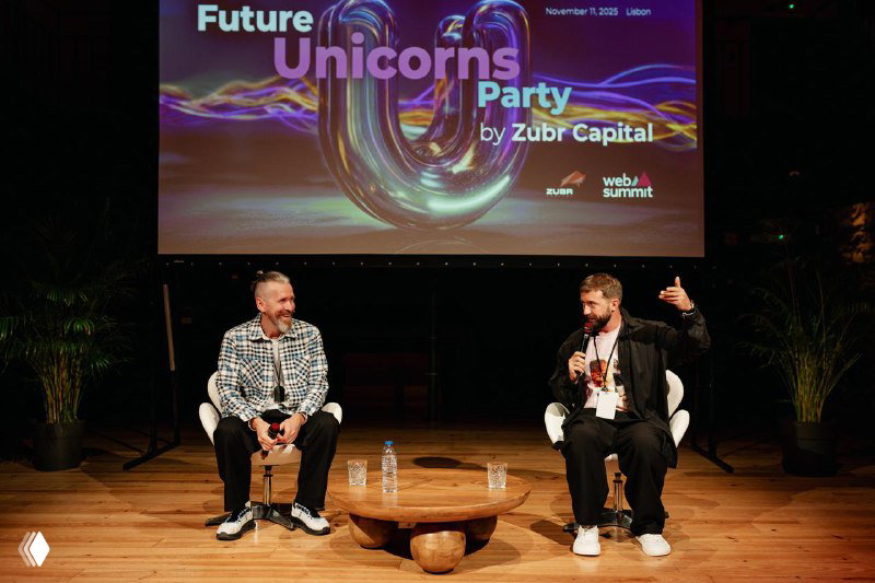 Two guests seated on stage in a panel format at the Future Unicorns Party 2025, wooden decor on stage and projection above them.