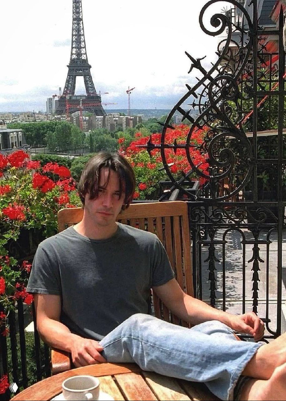 Мужчина в футболке и джинсах сидит на террасе с чашкой кофе, вдалеке видна Эйфелева башня и клумбы с цветами — спокойный повседневный кадр.