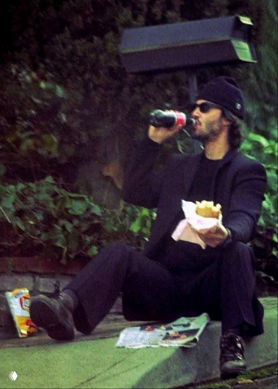 Кадр мужчины в тёмной одежде на улице, он сидит на бордюре с фастфудом и напитком, расслабленная уличная сцена с зеленью на заднем плане.
