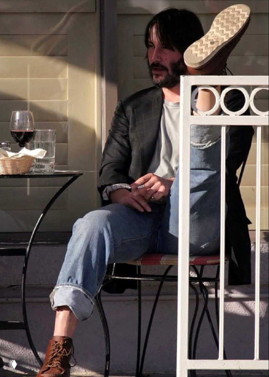 Человек в пиджаке и джинсах расслабленно сидит на веранде кафе с бокалом вина, непринуждённый момент из обычной жизни за столиком на свежем воздухе.