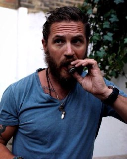 Седоволосый мужчина с густой бородой затягивается сигарой в садовом или уличном окружении, сцену пронизывает спокойствие.