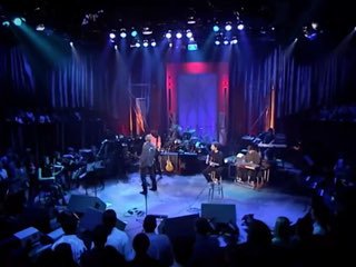 Живая сцена: солист в центре микрофона, группа музыкантов за ним и рассеивающаяся публика в затемнённом зале под синим светом.