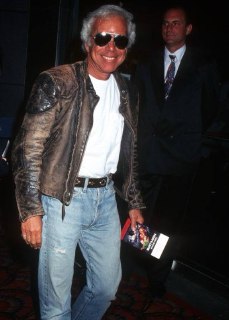 Улыбчивый зрелый мужчина в поношенной кожаной куртке и джинсах выходит в свет; кадр передаёт свободный стиль уверенного возраста.