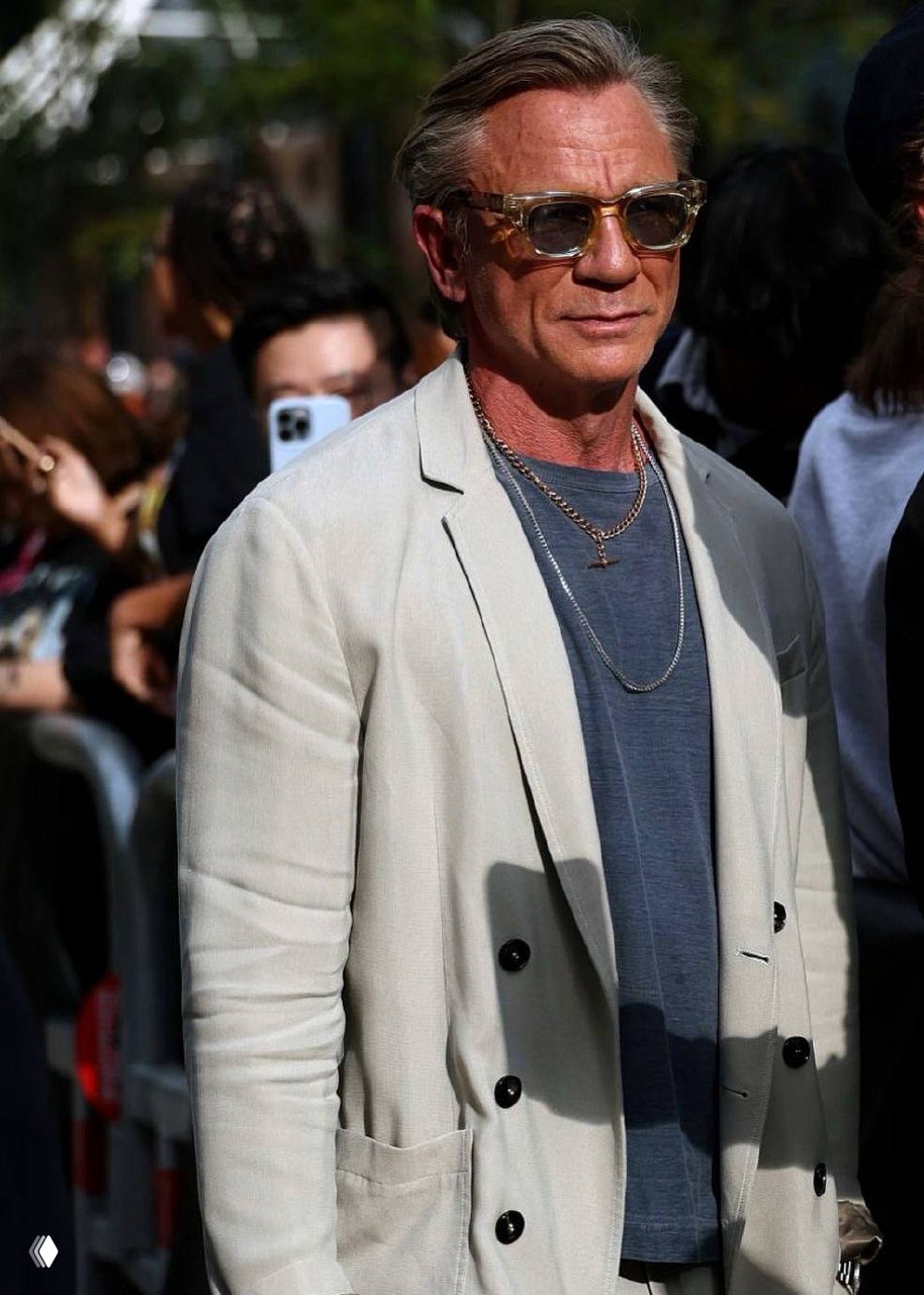 Зрелый мужчина в светлом двубортном пиджаке и солнцезащитных очках идёт по людной улице; сочетание небрежной элегантности и мужественности.