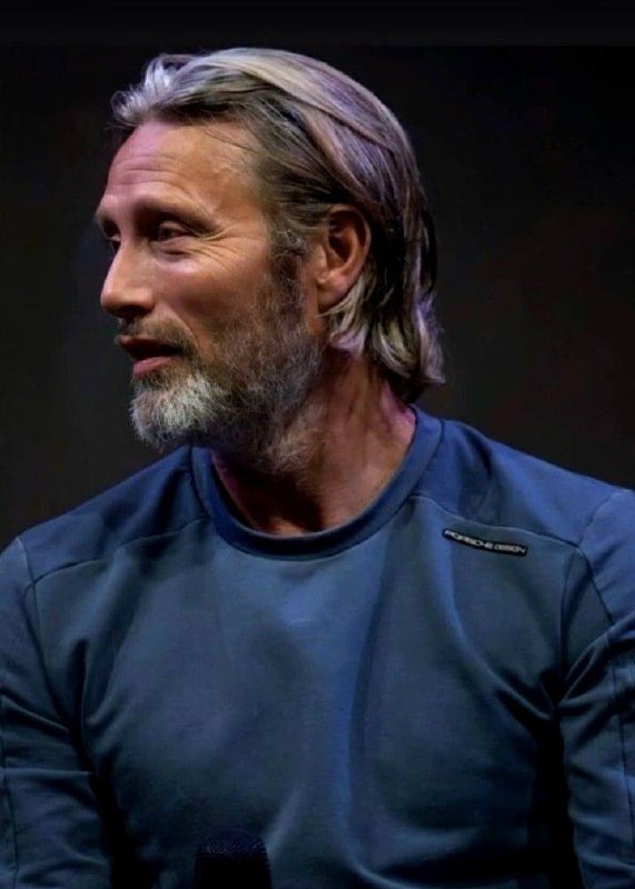 Портрет зрелого мужчины в повседневной одежде на сцене; седеющие волосы и спокойный профиль создают образ опыта и сдержанности.