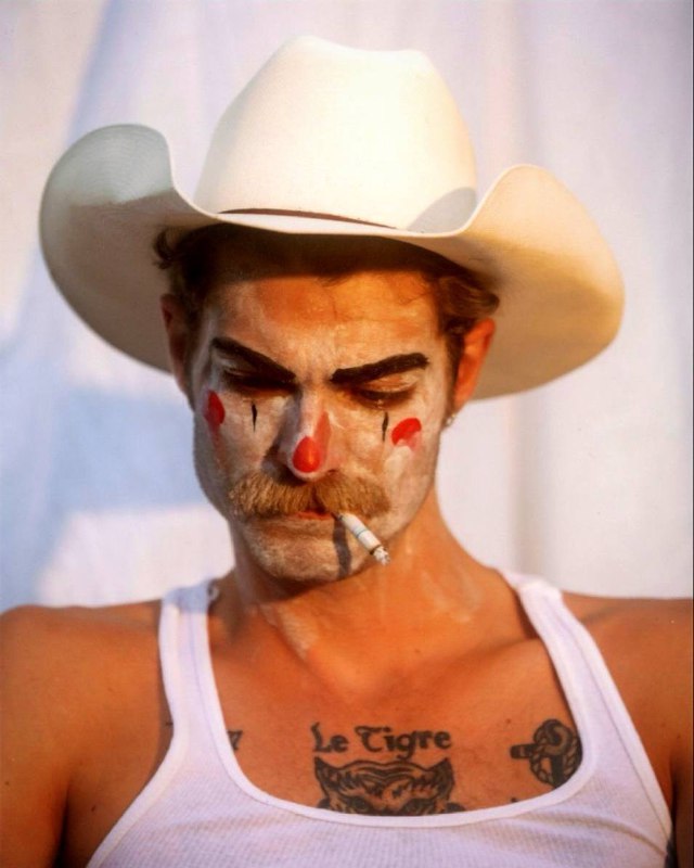 Мужчина в ковбойской шляпе с клоунским макияжем и сигаретой; фотография передаёт театральную и грубую эстетику портрета.