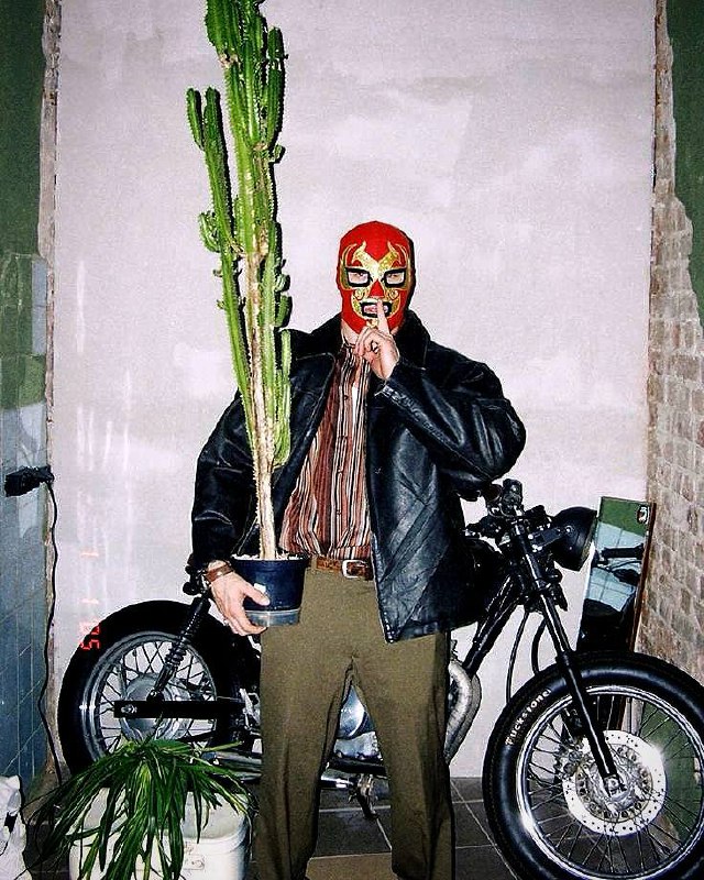 Человек в маске борца с кактусом в руках на фоне мотоцикла; уличный кадр с элементами сюрреализма и креативной эстетики.