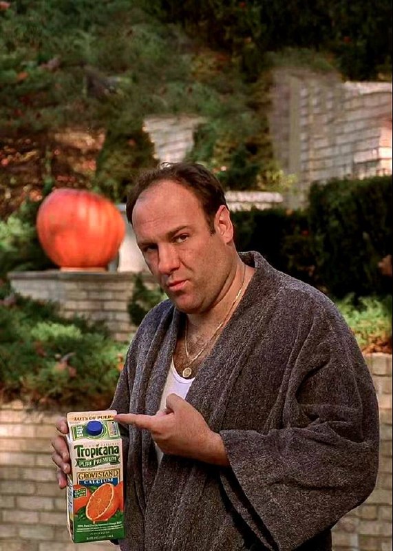 Тони Сопрано (Джеймс Гальдонфини) стоит в саду с коробкой сока в руке, в кадре виден задник с большим яблоком — бытовой и неожиданный образ персонажа.
