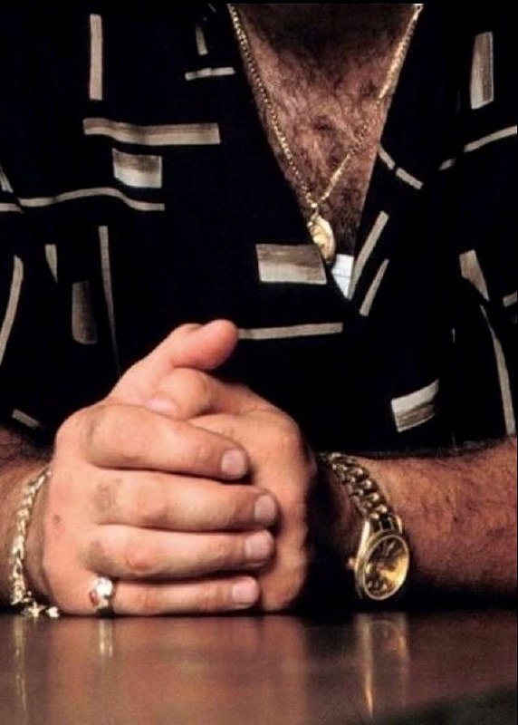 Крупный план рук Тони с кольцами, золотыми браслетами и сложенными руками на столе — акцент на аксессуарах и характерных мужских жестах персонажа.