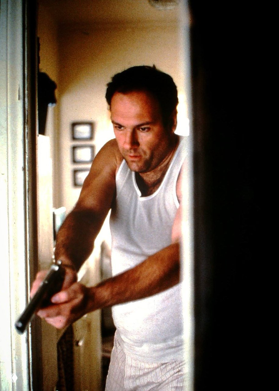 Тони Сопрано в майке целится из огнестрельного оружия в коридоре — напряжённый драматический кадр без графических деталей, подчёркивающий опасность сцены.