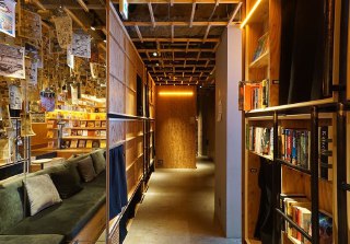 Длинный коридор с деревянными стеллажами и подсветкой, ведущий к капсулам в гостинице Book and Bed в Токио.