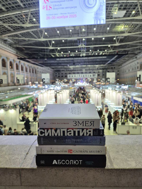 Стопка купленных книг на перилах с видом на ряды non/fiction: на переднем плане «Змея», «Симпатия» и «Абсолют», за ними — людные коридоры стендов.