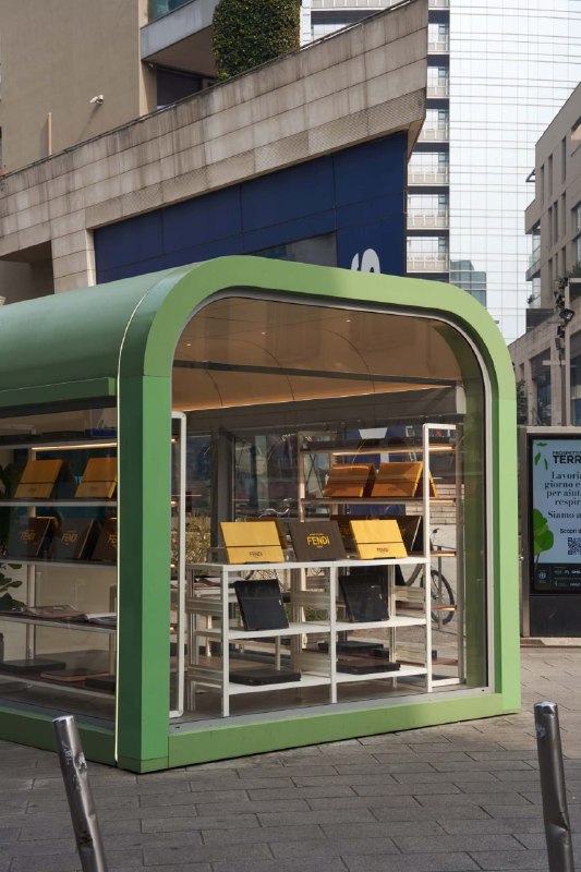 Другая ракурсная фотография зелёной модульной точки продаж с металлической конструкцией и витринными полками, заполненными печатной продукцией.