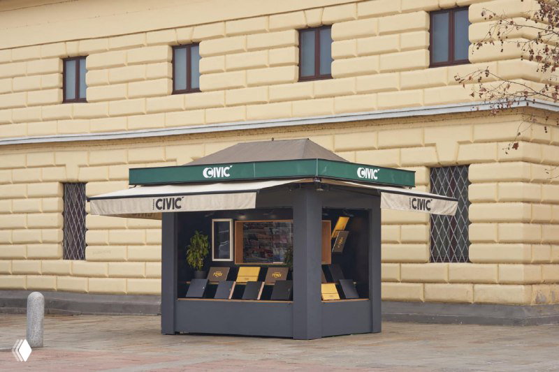 Квадратная открытая киосковая конструкция с табличками и полками, на которых выставлены книги и журналы — пример формата pop‑up для книгораспространения.