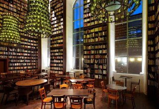 Вечерний вид библиотеки отеля B2: деревянные столы, высокие окна и стены, полностью занятые книгами на нескольких уровнях.