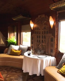 Обеденная зона в сьюте: круглый стол, уютные диваны, сервировка и декоративная панель у окна в элегантном интерьере поезда.