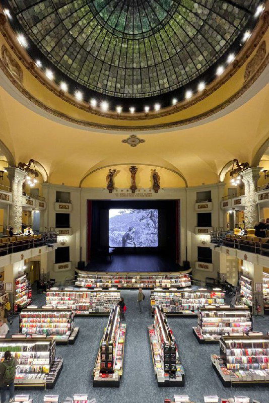 Вид сверху на центральный проход Giunti Odeon: симметричные книжные стеллажи, лестницы и большой экран в глубине зала, интерьер в классическом стиле.