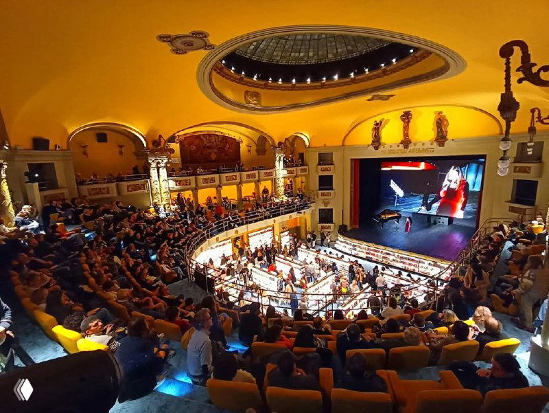 Широкий панорамный вид на переполненный зал Giunti Odeon во время мероприятия: зрители, сценическое освещение и окружение книжных стеллажей.