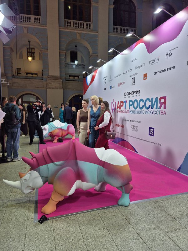 Посетители на фоне цветных надувных или скульптурных носорогов на розовой подложке у фотозоны ярмарки, люди позируют.